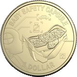 A gold-colored circular coin or medallion with embossed safety-themed graphics and text reading 'BABY SAFETY CAPSULE' around the edge and '1 DOLLAR' at the bottom. The center shows a baby capsule and infant figure in a stroller, with small icons of a car, bottle, and light bulb.